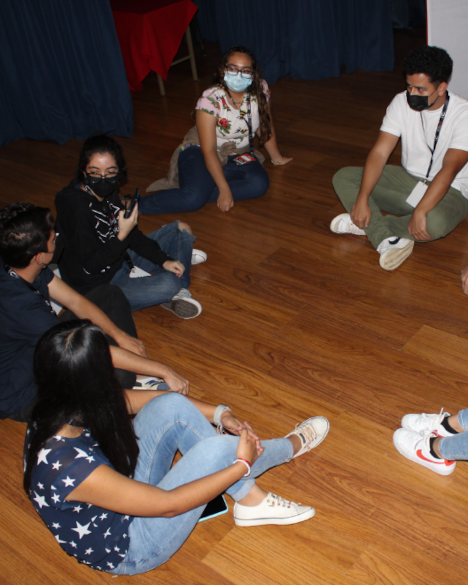 Grupo de estudiantes sentados en el piso, en círculo y hablando.